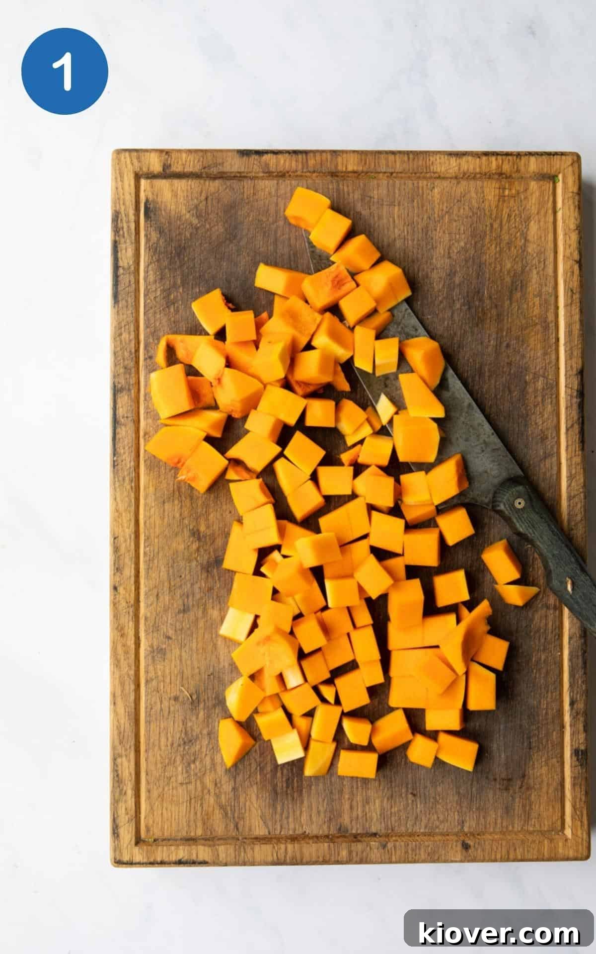 Butternut squash cut into cubes laid out on a wooden chopping board with a knife positioned on a white marble background.