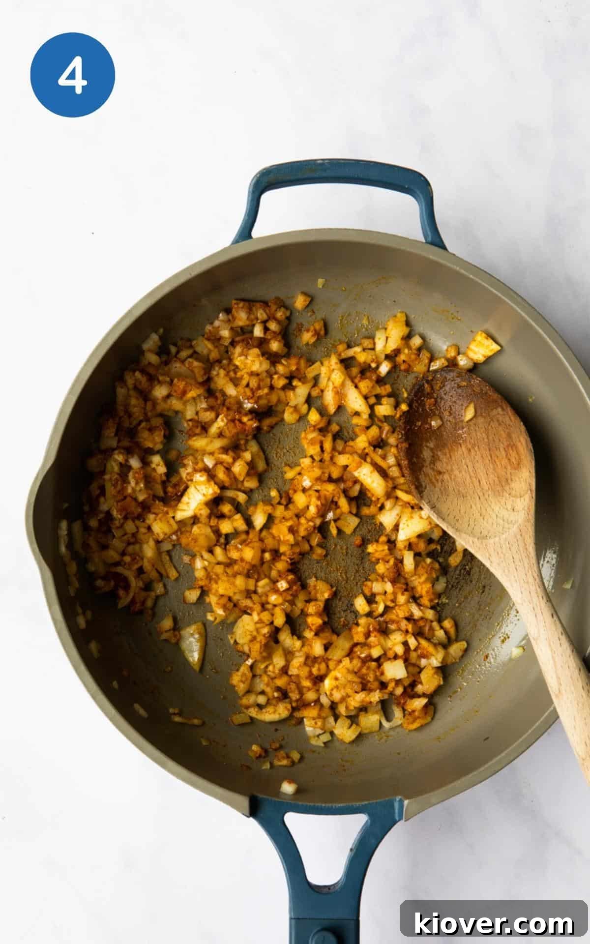 Onion being cooked with spices in a large non stick frying pan with wooden spoon. White marble background. 