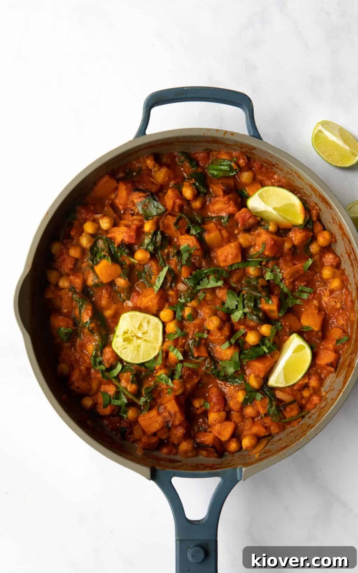 Butternut Squash and Chickpea curry in a large non-stick frying pan topped with lime wedges and chopped coriander on a white marble background. 