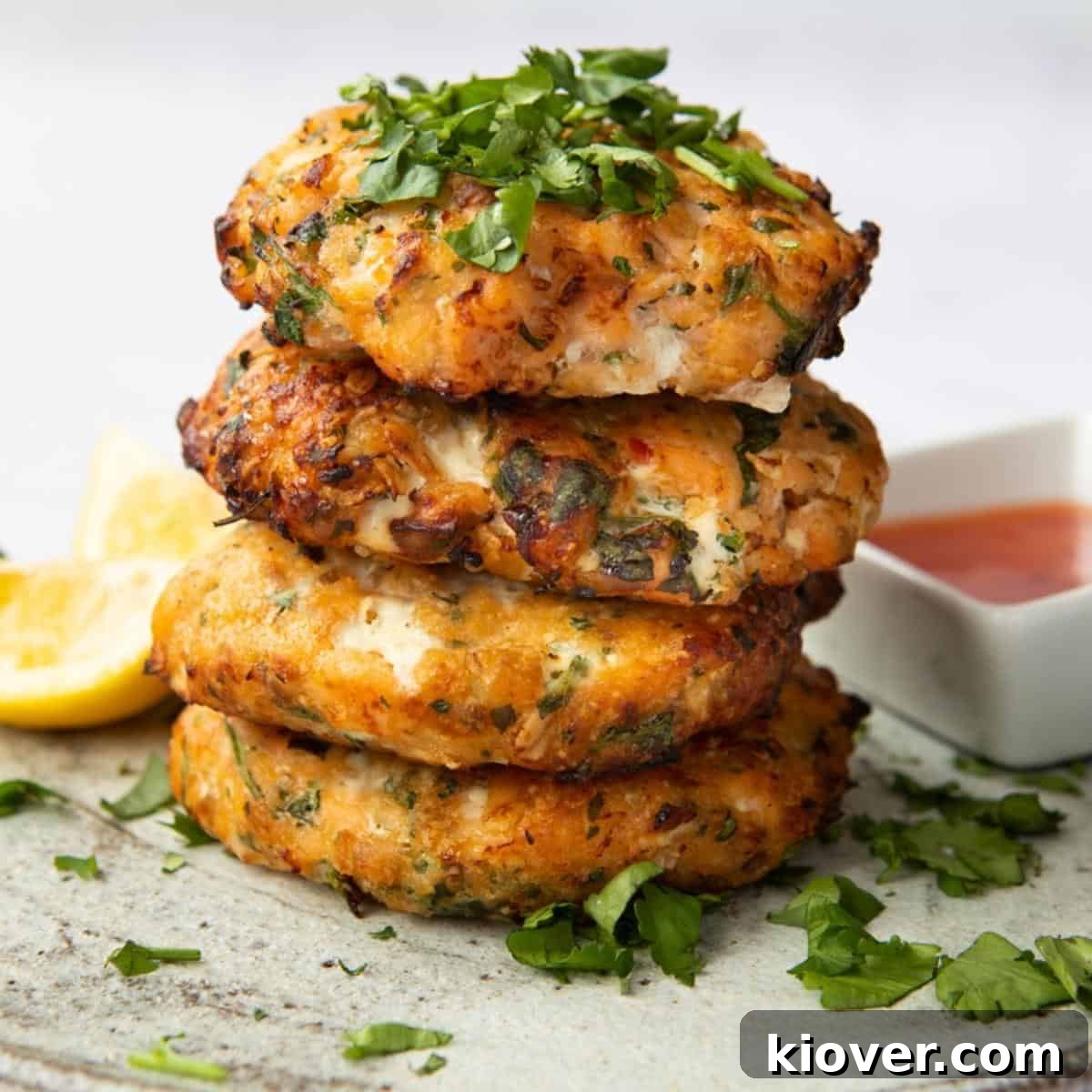 4 Sweet Chilli Salmon Fishcakes stacked on a plate topped with coriander. Lemon wedges and a dish of sweet chilli sauce are blurred in the background. 