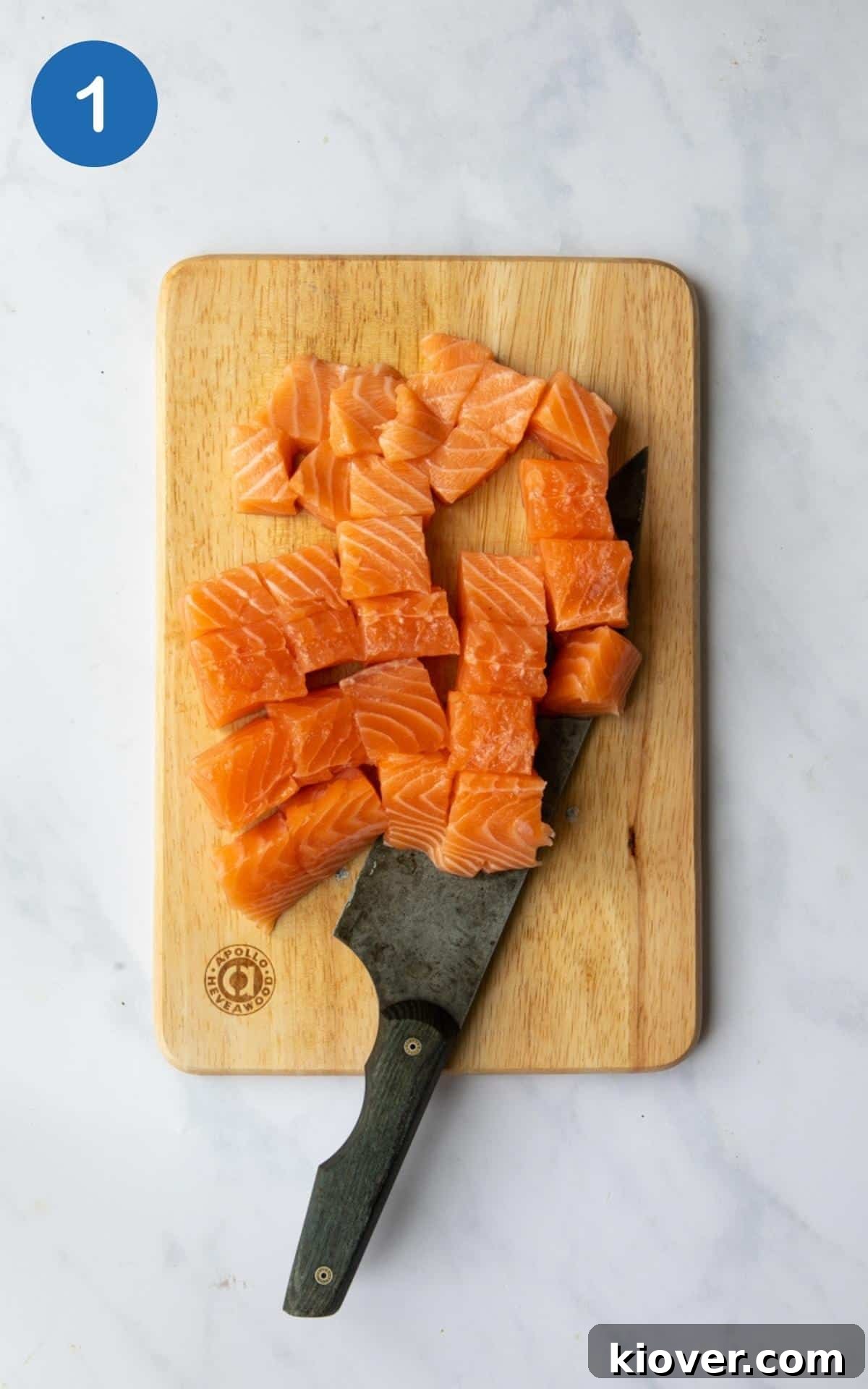 Salmon cut into chunks on a wooden chopping board with knife on a white marble background.