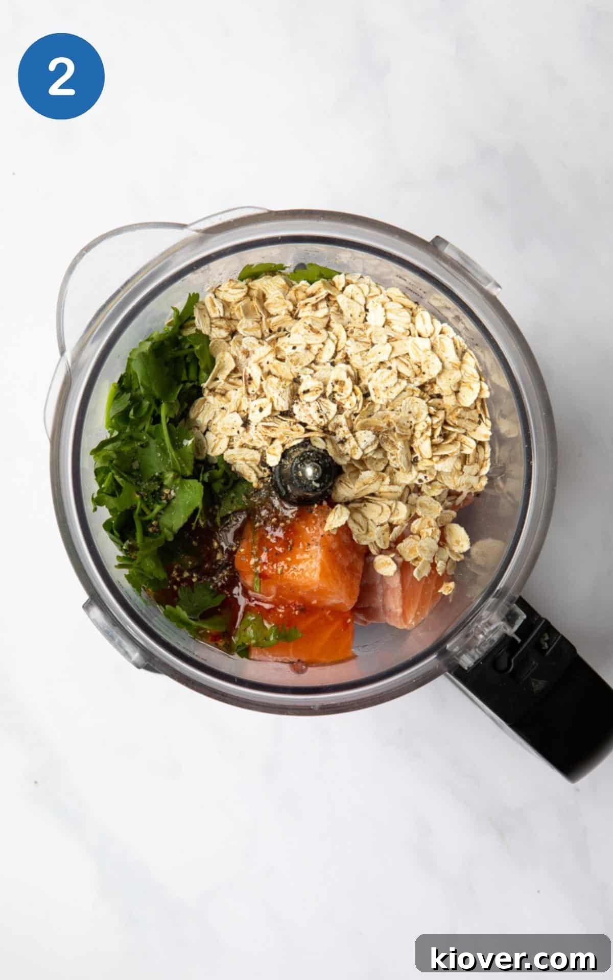 A food processor showing pre-blended ingredients: salmon, oats, coriander, lemon juice and sweet chilli sauce in it. Presented on a white marble background.