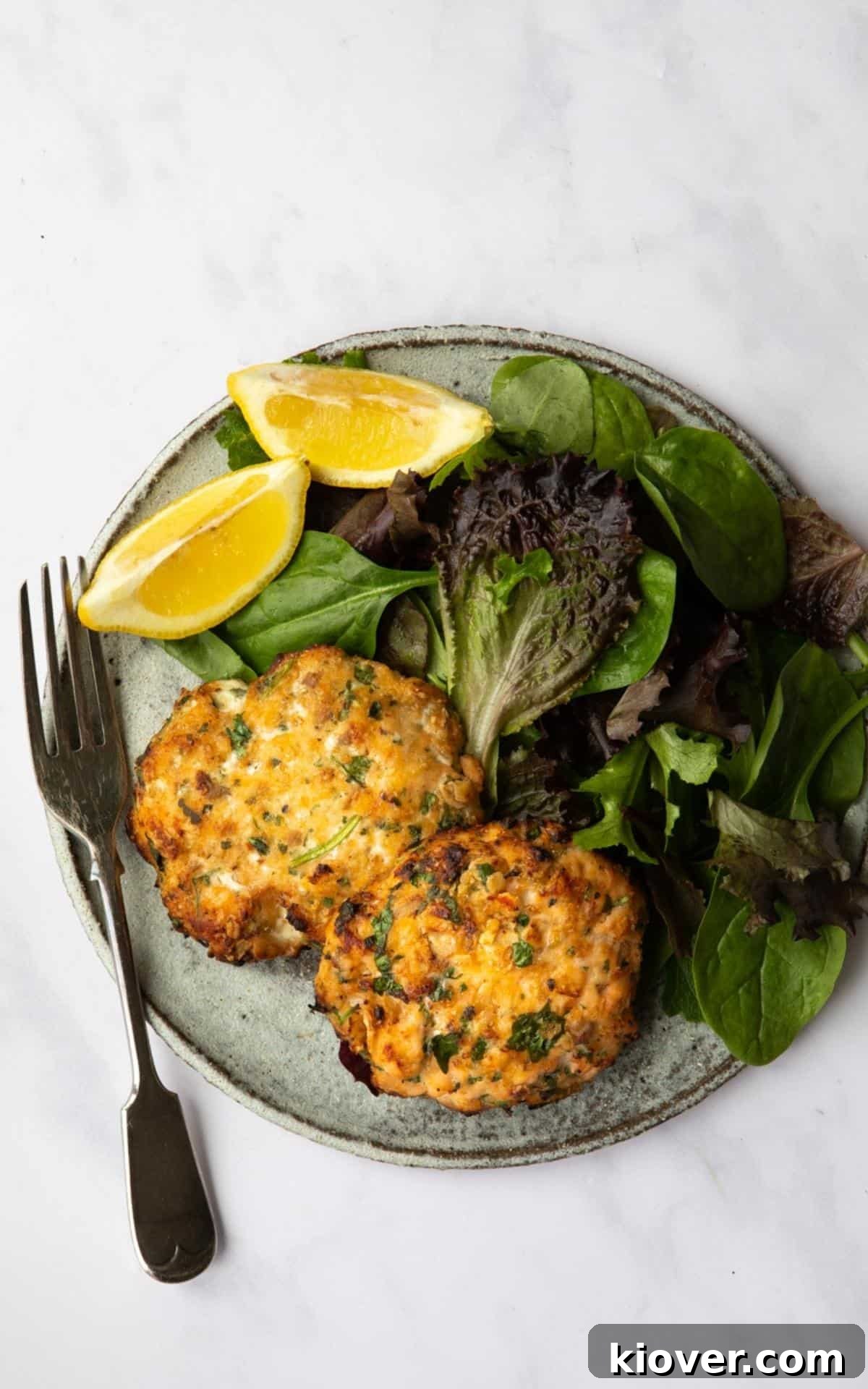 Sweet Chilli Salmon Fishcakes served on a plate with salad leaves and lemon wedges. Presented on a white marble background.