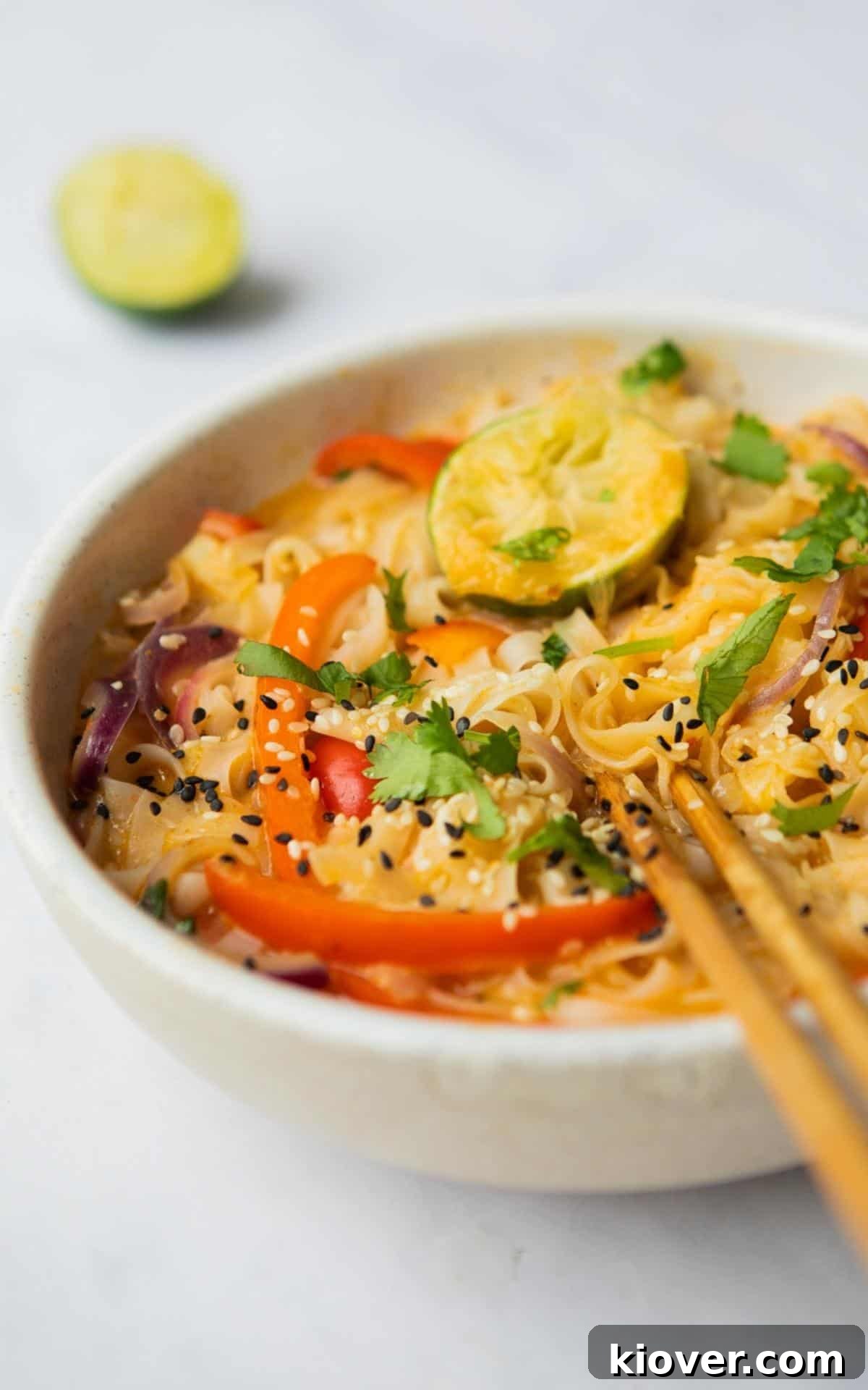 Thai Curry Noodle Soup Recipe — Creamy Thai Coconut Noodle Bowl 8 Close up image of Thai Curry Noodle Soup in a bowl with chopsticks. Blurred image on lime wedge in the background. Displayed on a blurred white marble background.