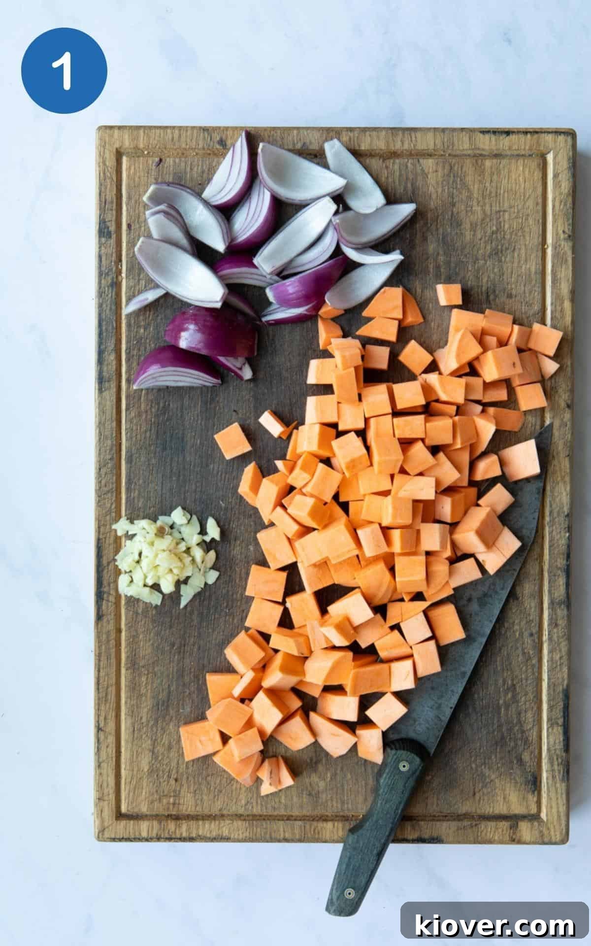 chopped sweet potato, finely sliced garlic and onion cut into chunks displayed on a wooden chopping board with knife. Board is displayed on a white marble background.