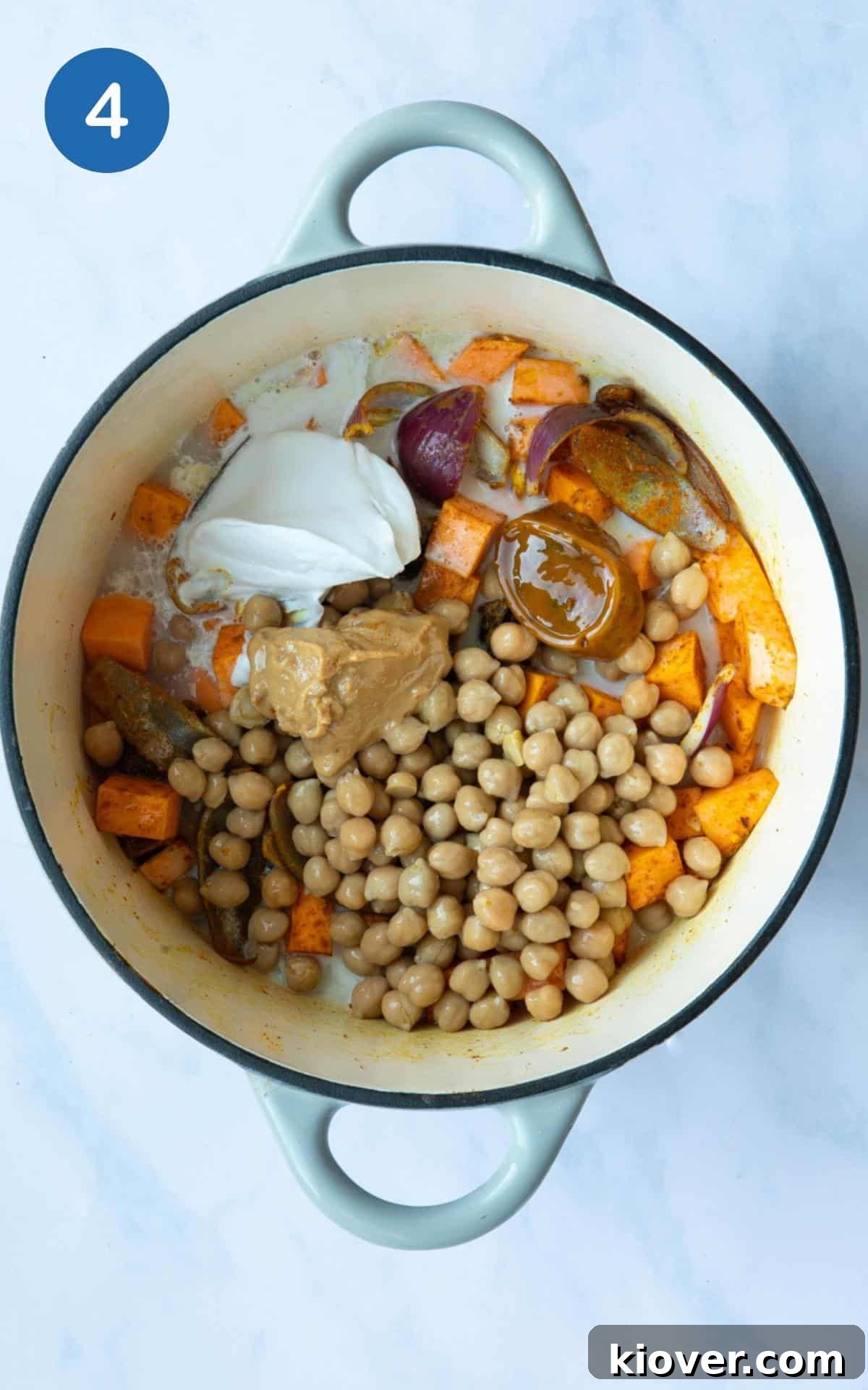 large pot displayed on white marble background with uncooked chickpeas, cubed sweet potato and onion, dollop of coconut milk, vegetable stock cube and peanut butter.