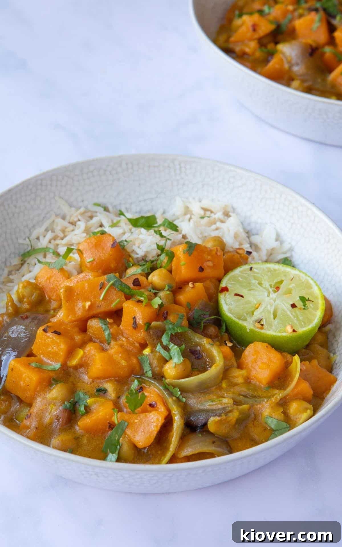 Sweet Potato Satay Curry served in a bowl with rice, topped with coriander and lime wedges. Bowl is displayed on a white marble background.