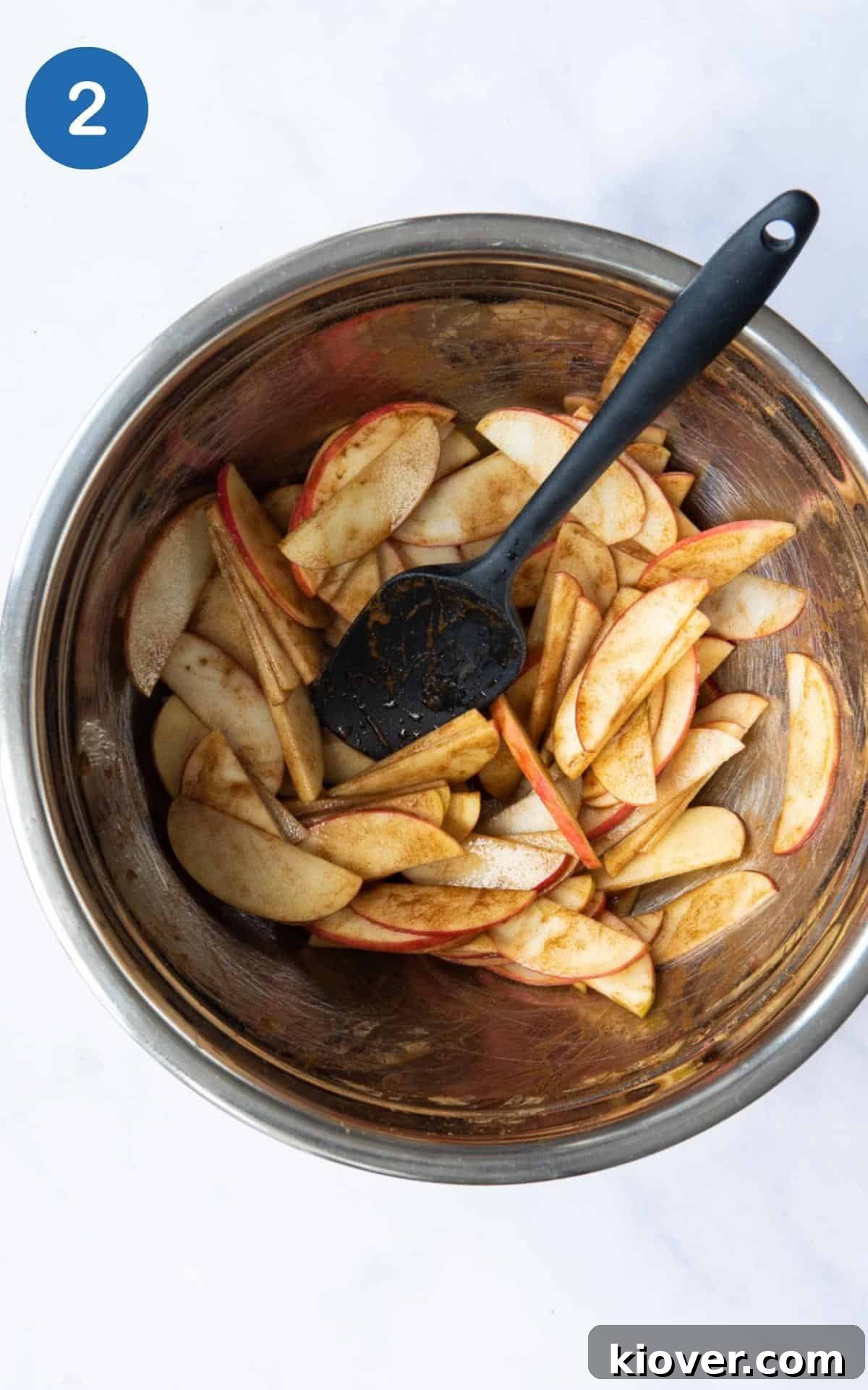 Apples and maple syrup being mixed together with a black spatula in a large silver mixing bowl.