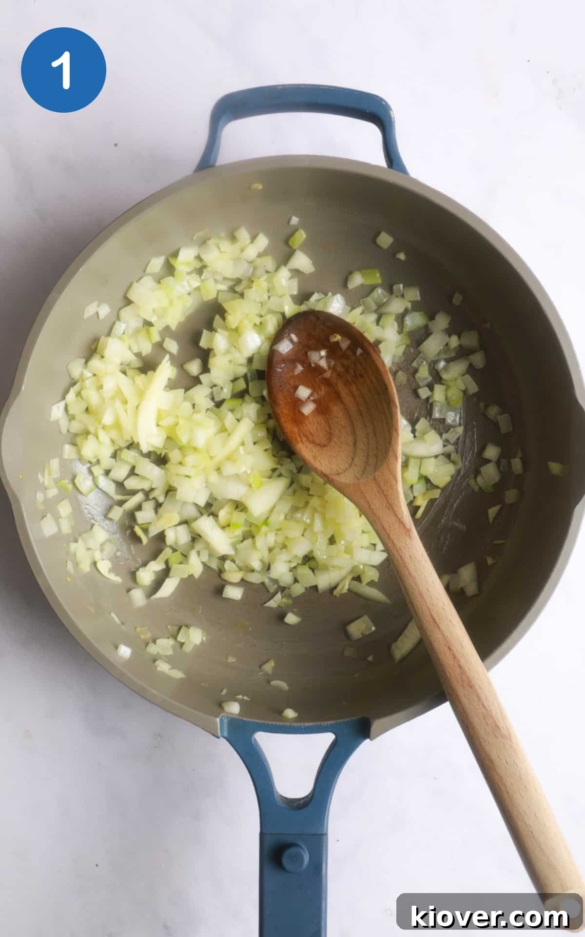onions being fried in large frying pan with wooden spoon. Pan is on white marble background.