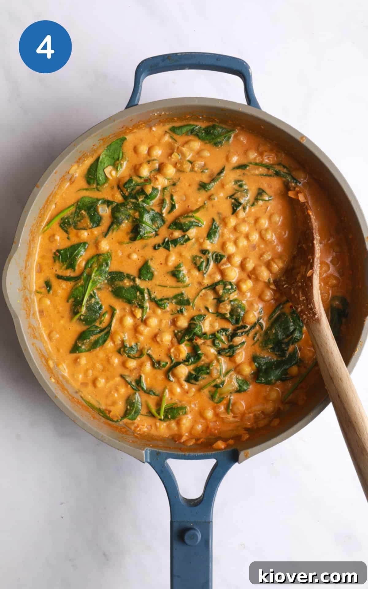 overhead shot of chickpea curry with wooden spoon in large frying pan. Pan is on white marble background.