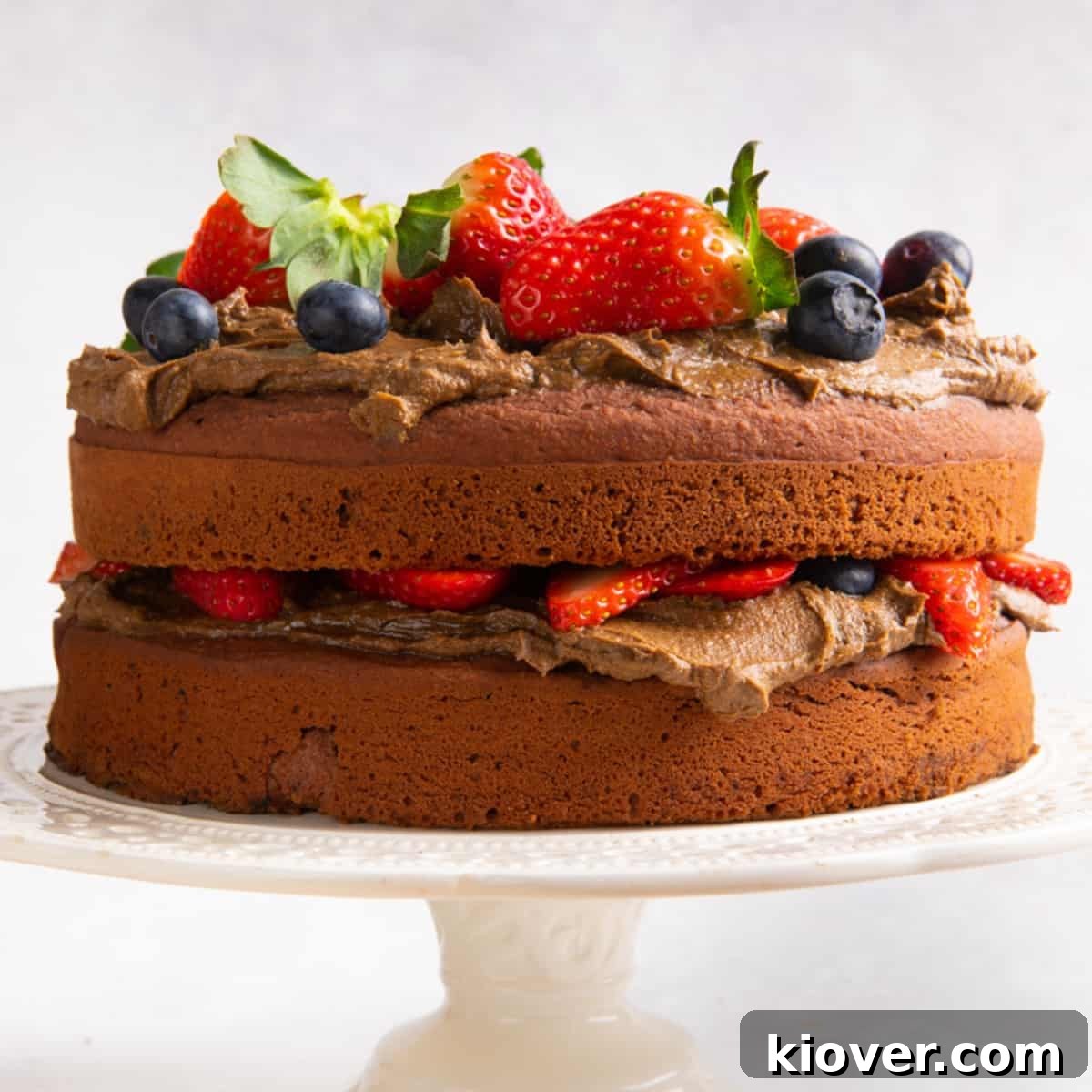 Chocolate cake topped with fresh berries on cream cake stand and white background.