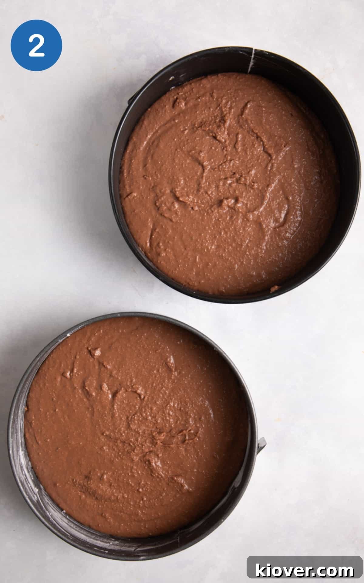 cake batter in 2 cake tins ready to be baked on white surface.
