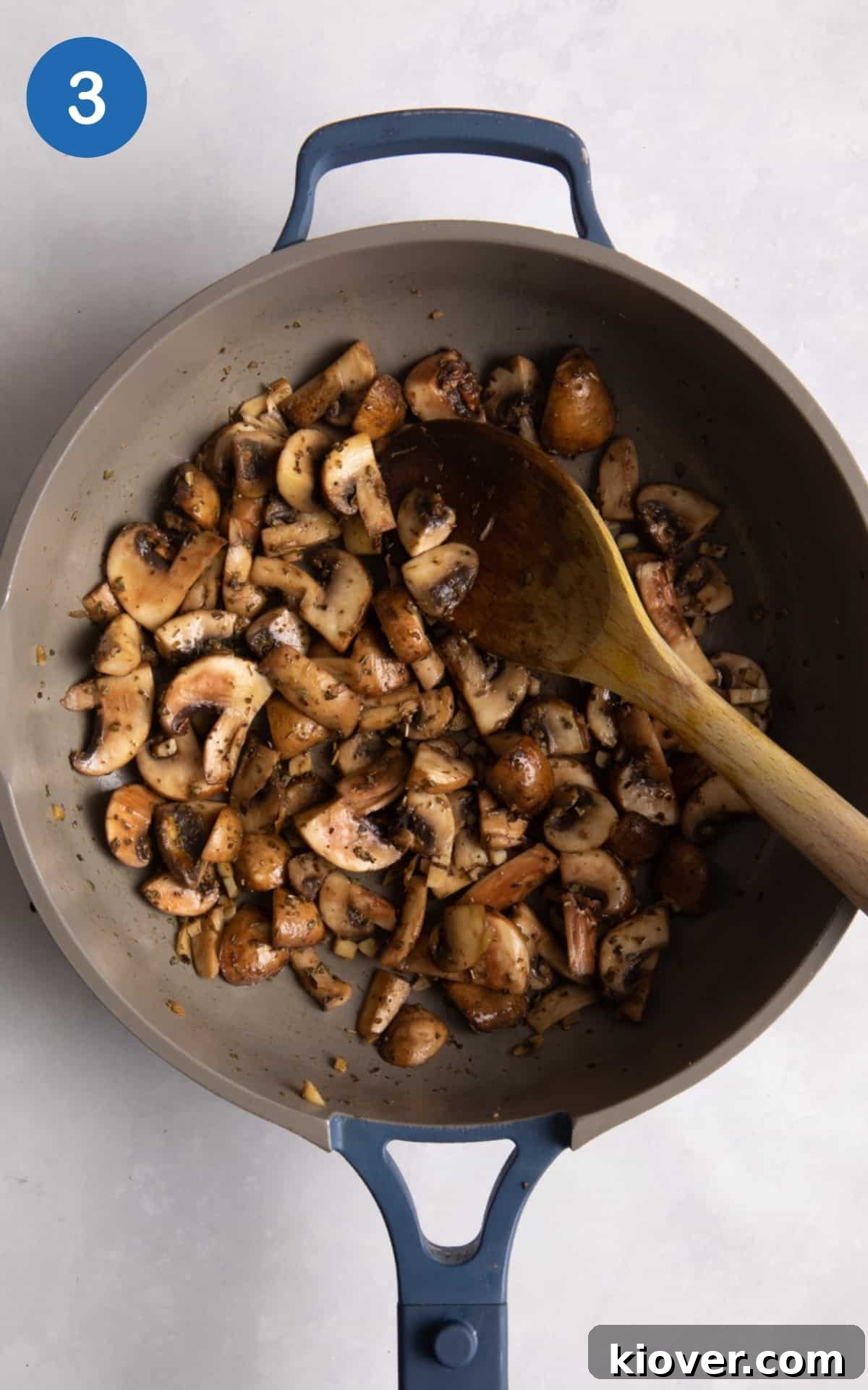 Garlic Butter Mushrooms on Toast with Herbs 6 cooked garlic mushrooms in blue frying pan with wooden spoon on white surface.
