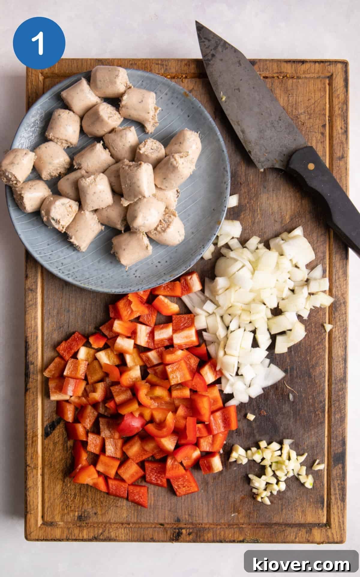 Chopped sausages, onion and red pepper on wooden chopping board.