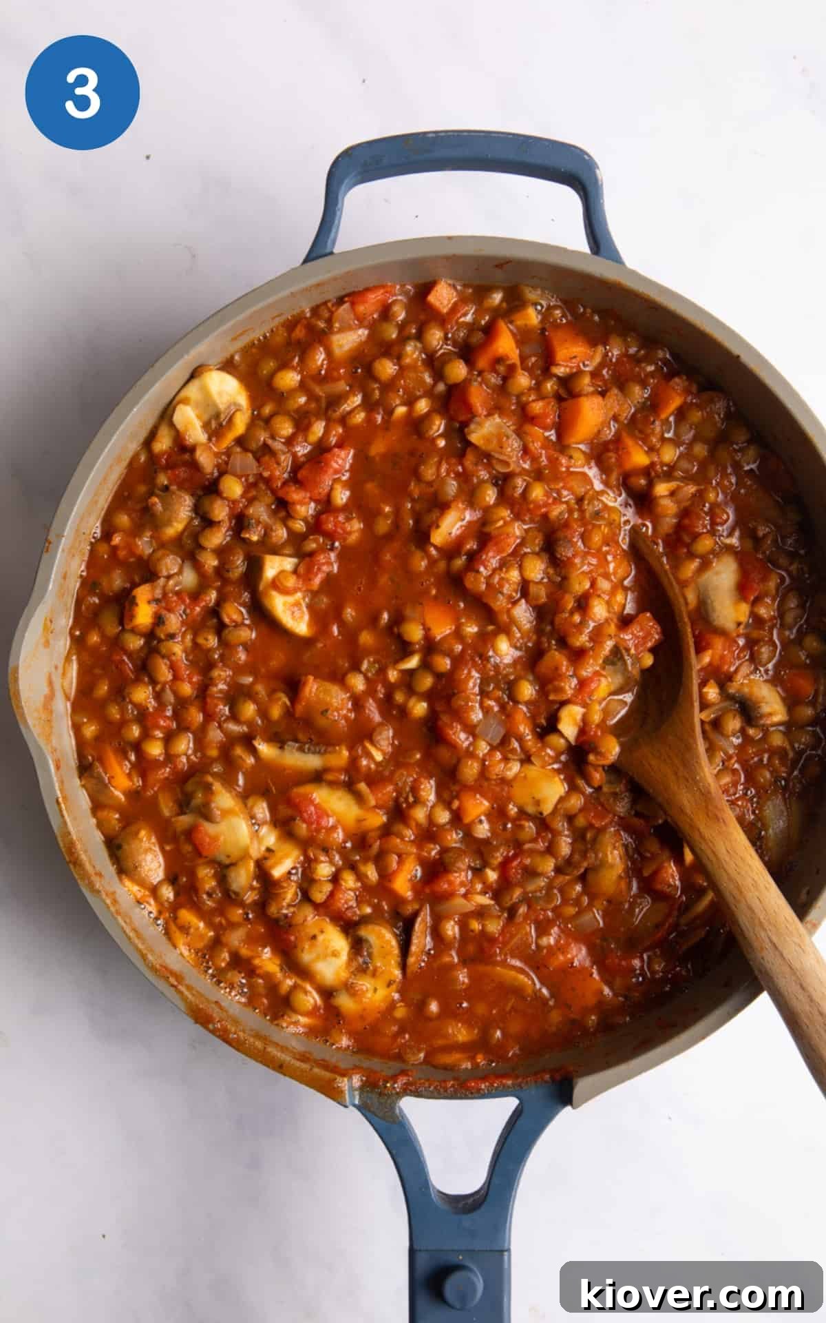 20 minute Lentil bolognese being cooked in a large frying pan with wooden spoon. Pan is displayed on white marble background. 