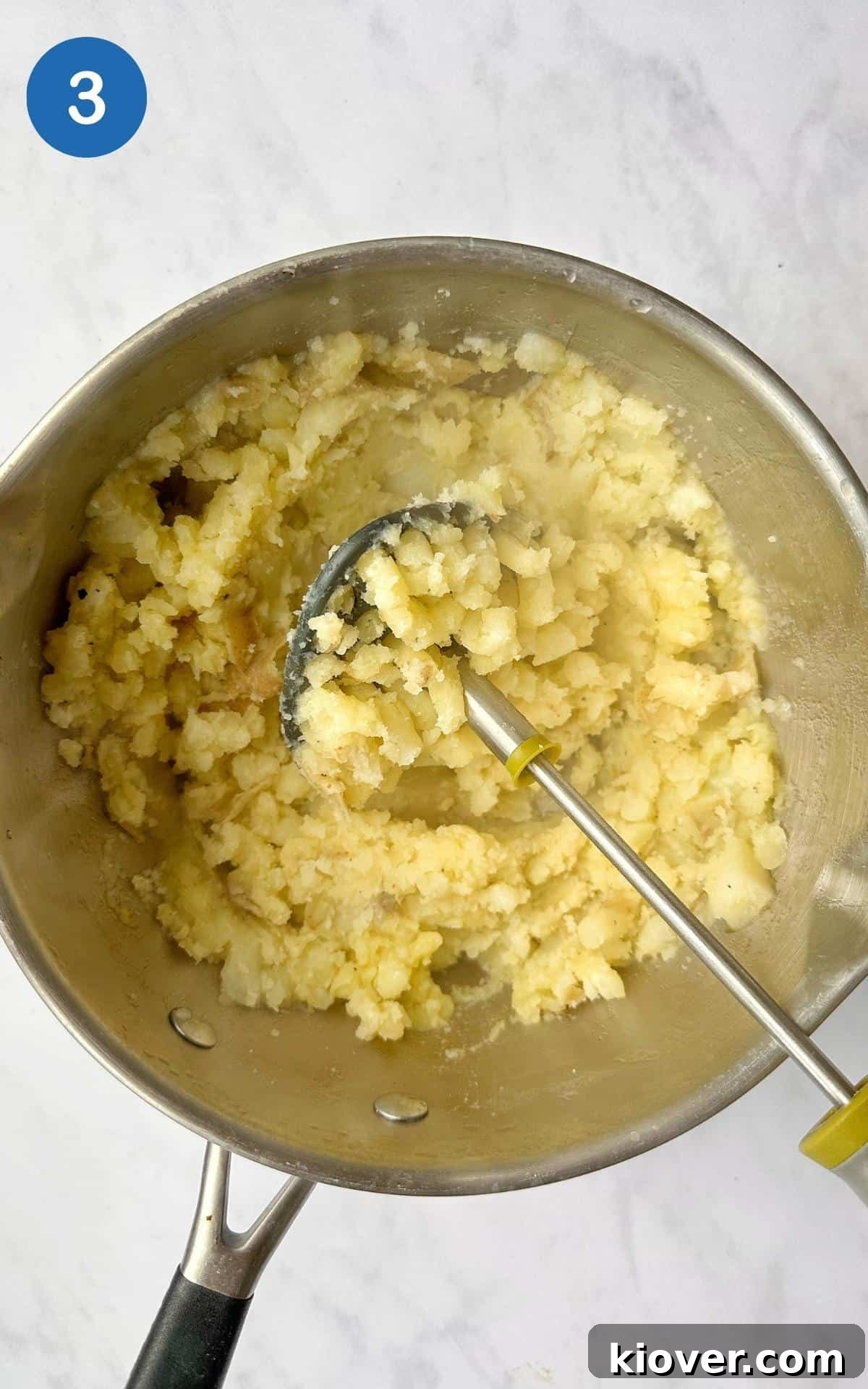 Potatoes being mashed in a saucepan. Saucepan is presented on white marble background.