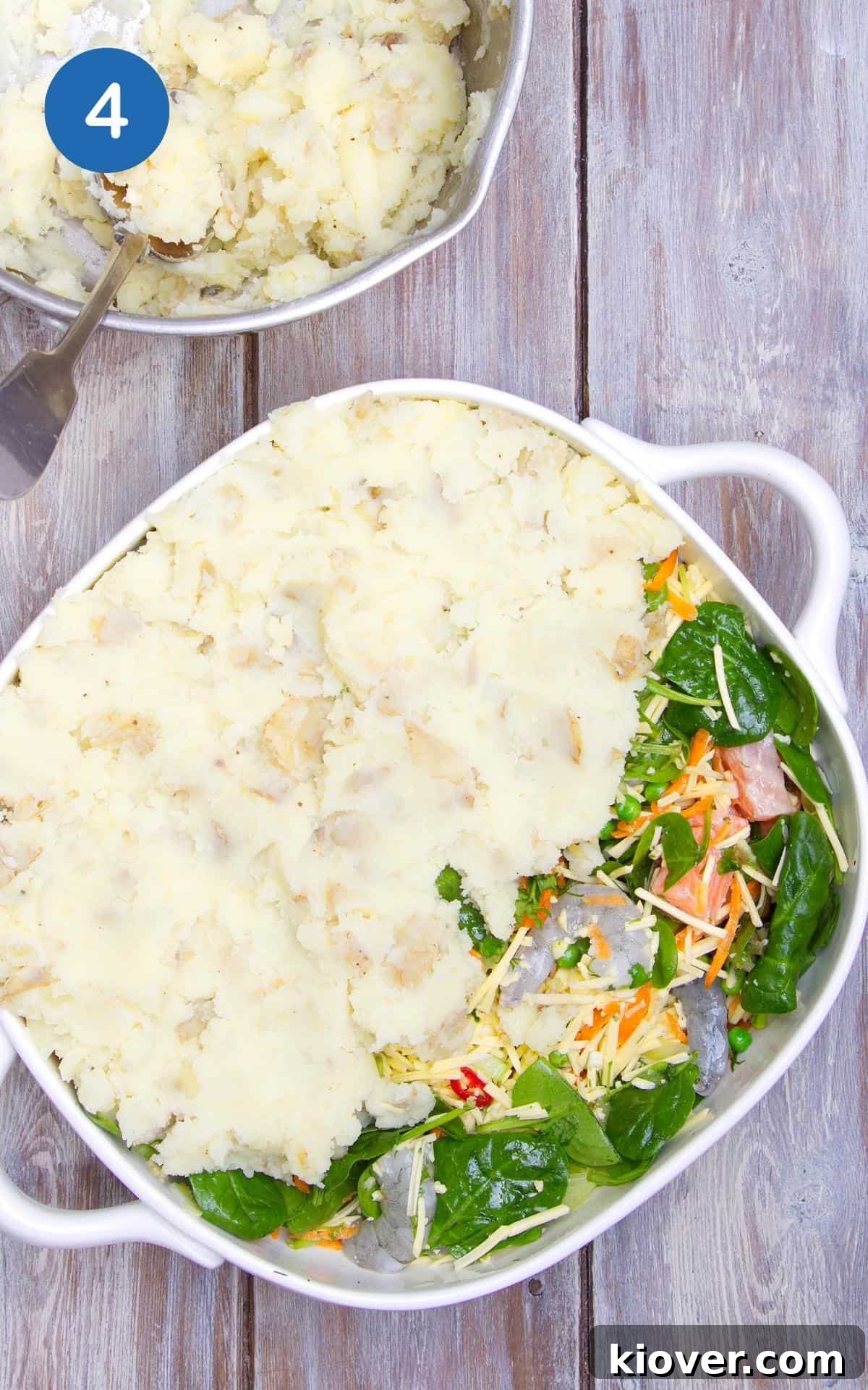 Mashed potato being put on top of the fish pie base mixture. Baking dish is presented on wooden table.
