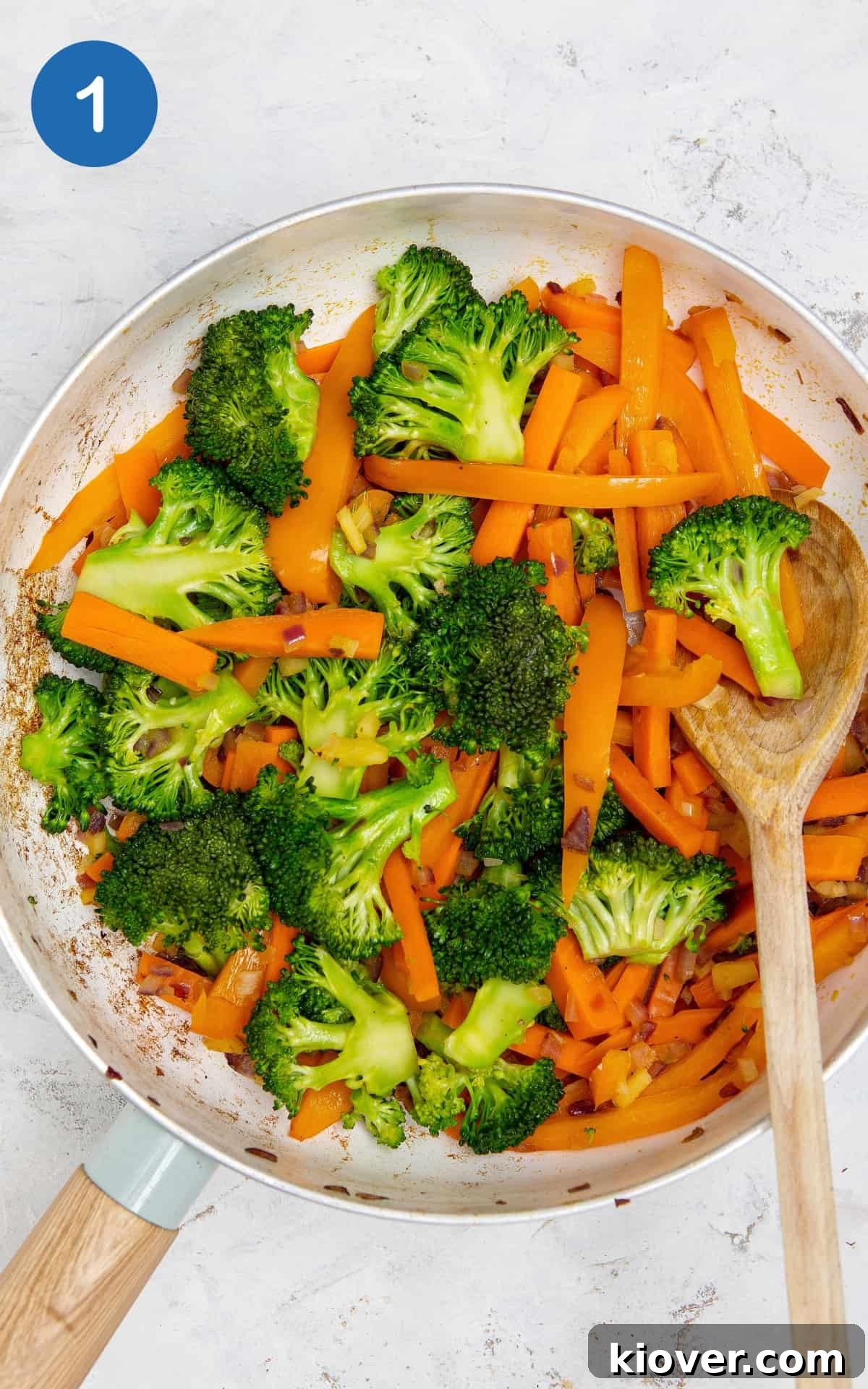 Broccoli and carrots in a frying pan with a wooden mixing spoon.