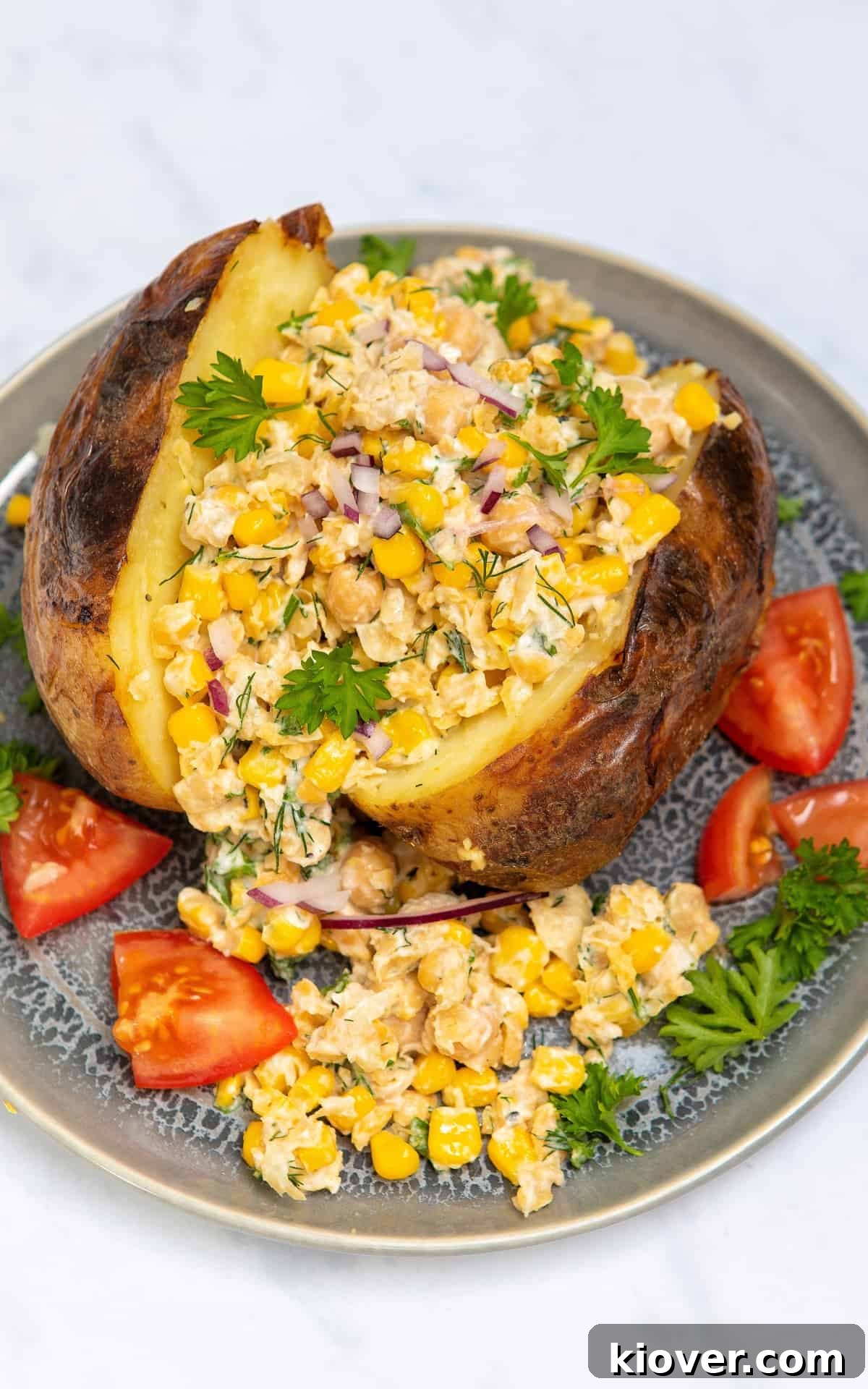 Chickpea and Tuna Salad Recipe for Meal Prep 6 chickpea mayo salad on a jacket potato surrounded by tomatoes and herbs on a blueish grey plate.