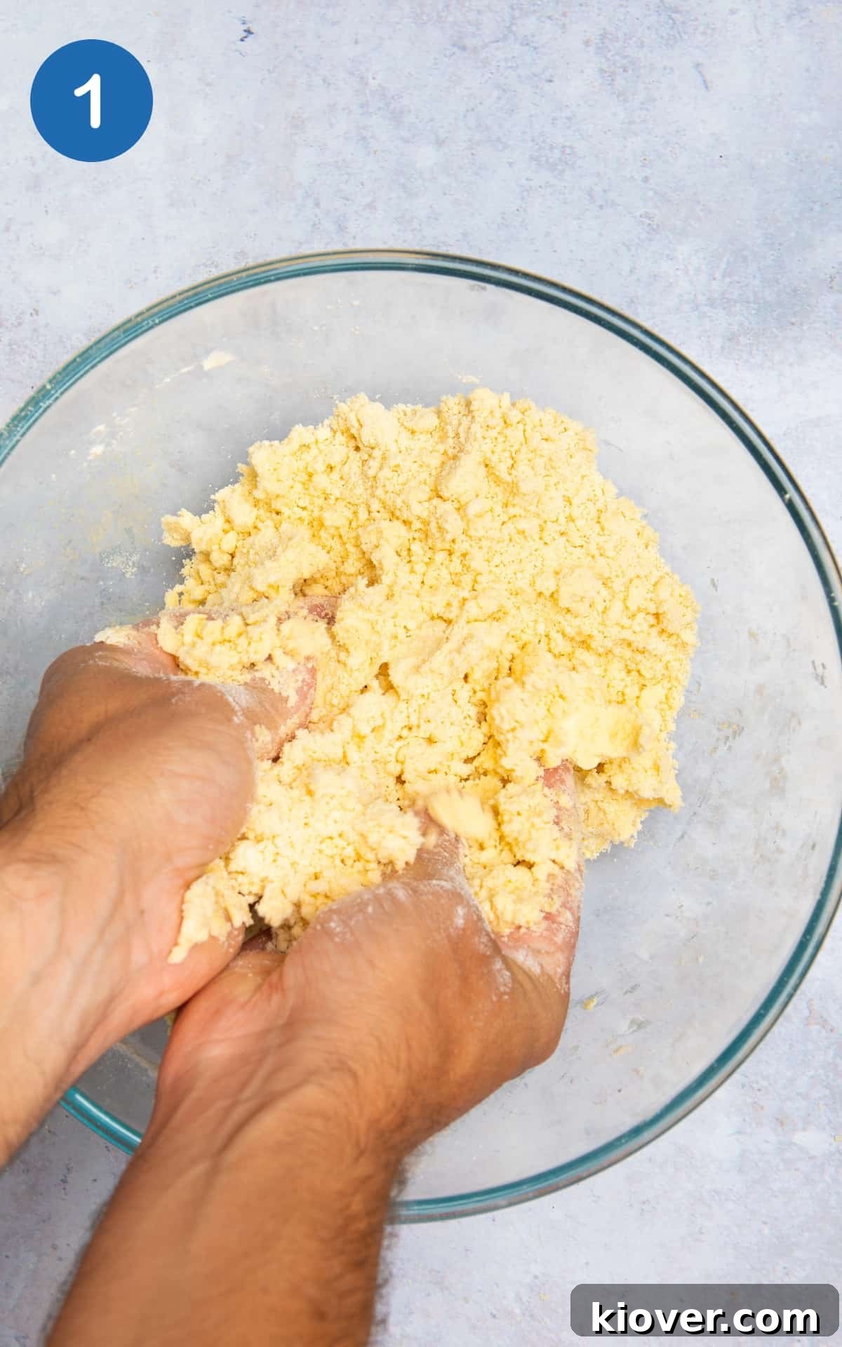 quiche pastry being crumbed with hands in transparent mixing bowl on grey surface.