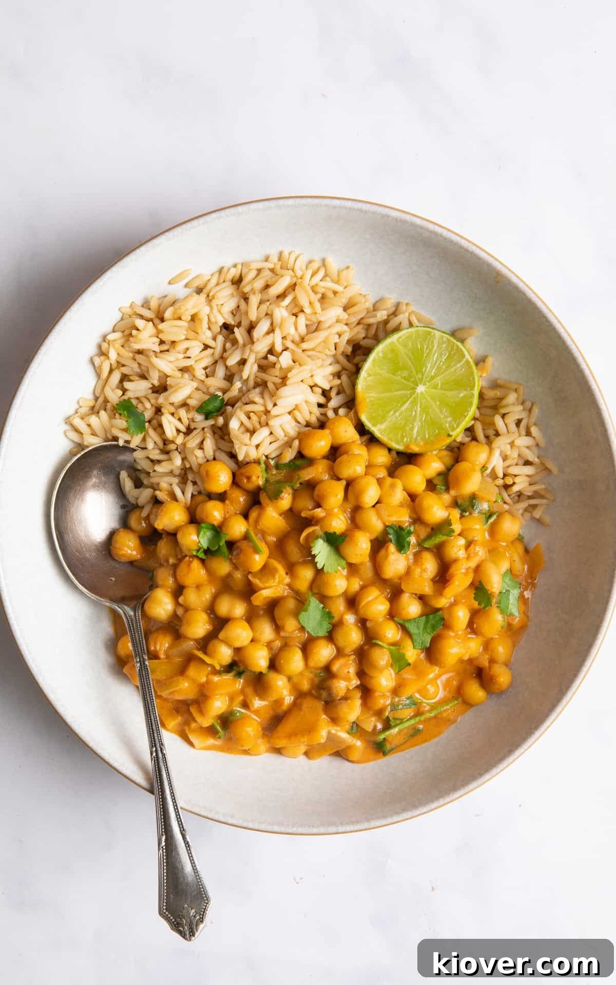 Curry in white bowl with rice and lime. There is also a silver spoon in the left hand side of the bowl. Bowl is presented on white marble background.