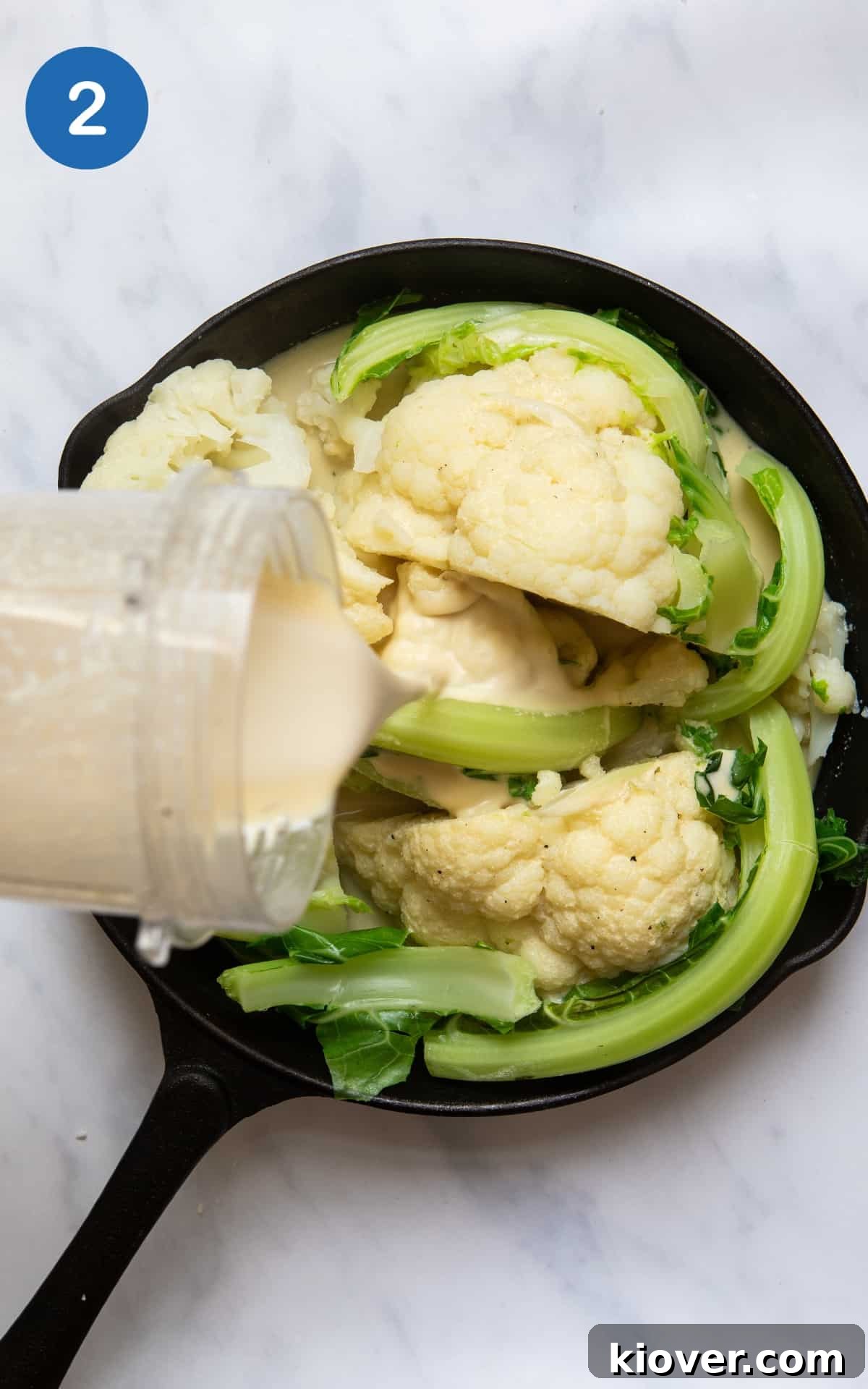 Cooked cauliflower in black skillet pan with sauce being poured over it. Skillet pan is on white marble background.