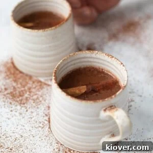a closeup of this homemade Christmas Hot Chocolate in two white coffee mugs garnished with cacao powder and cinnamon sticks.