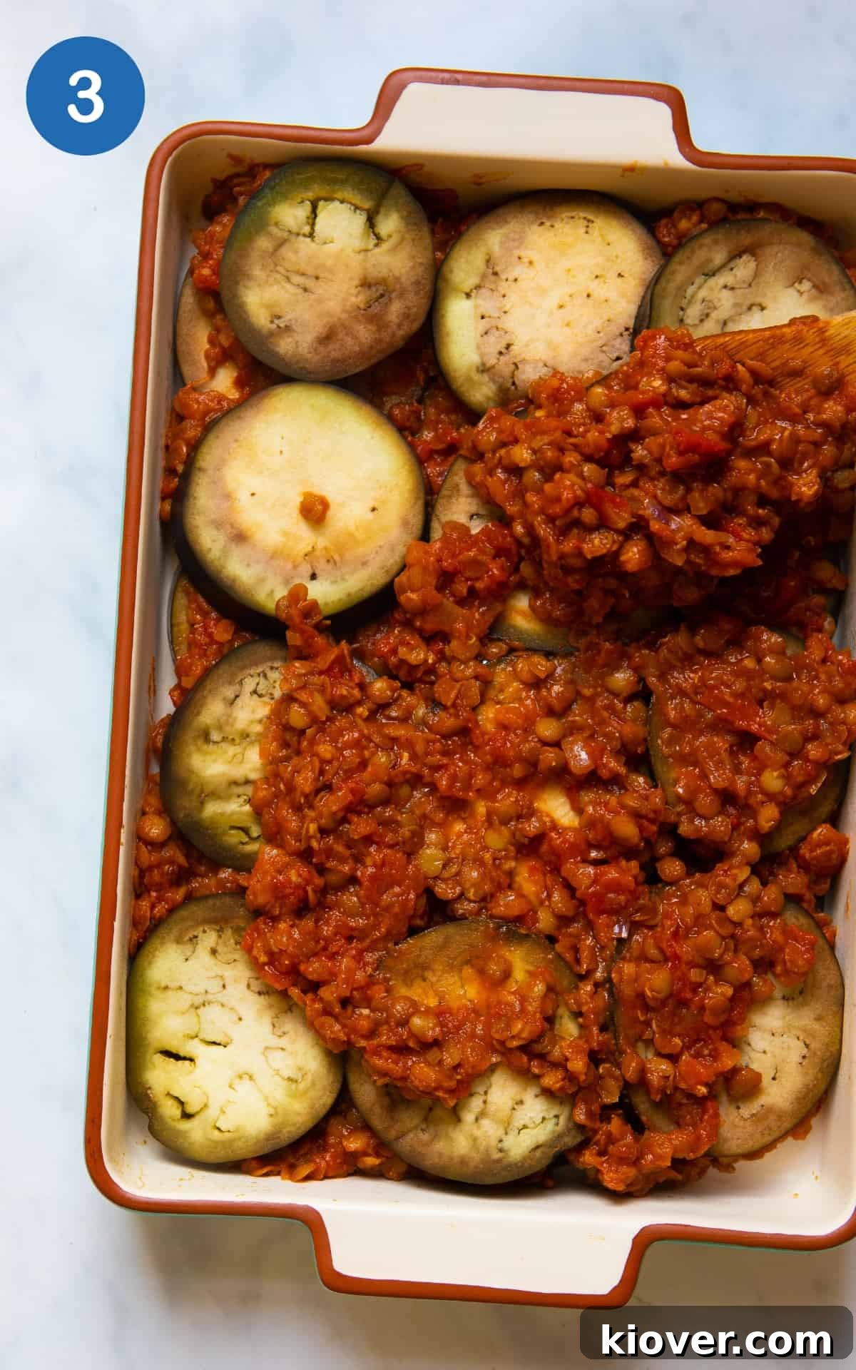 Creamy Baked Eggplant Casserole with Garlic and Herbs 6 Aubergine's being layered with lentil mix in a large baking dish. Baking dish is on white marble background.