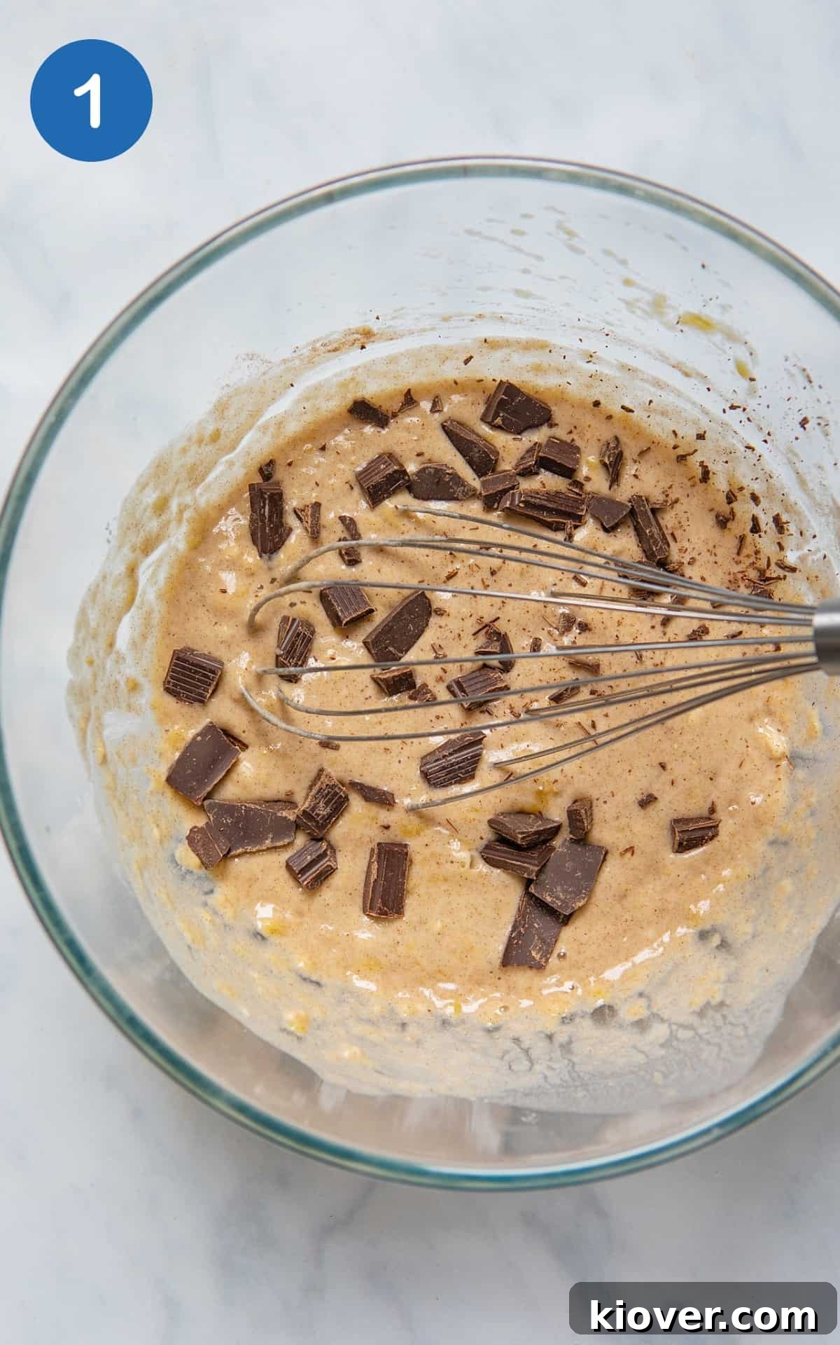 banana pancake batter mixed in a glass bowl on a white marble background with a whisk showing.