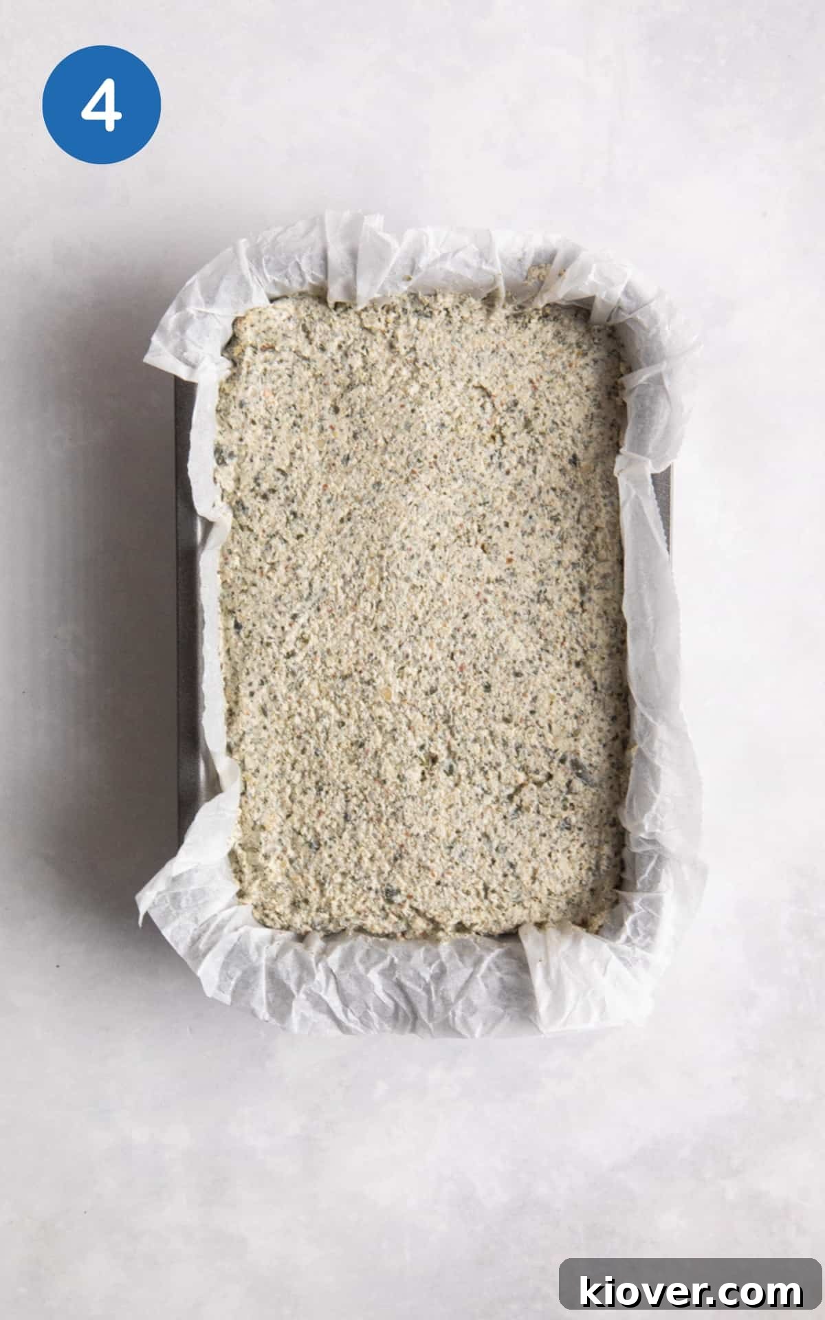 uncooked seeded bread in loaf baking tin with parchment paper on white surface.